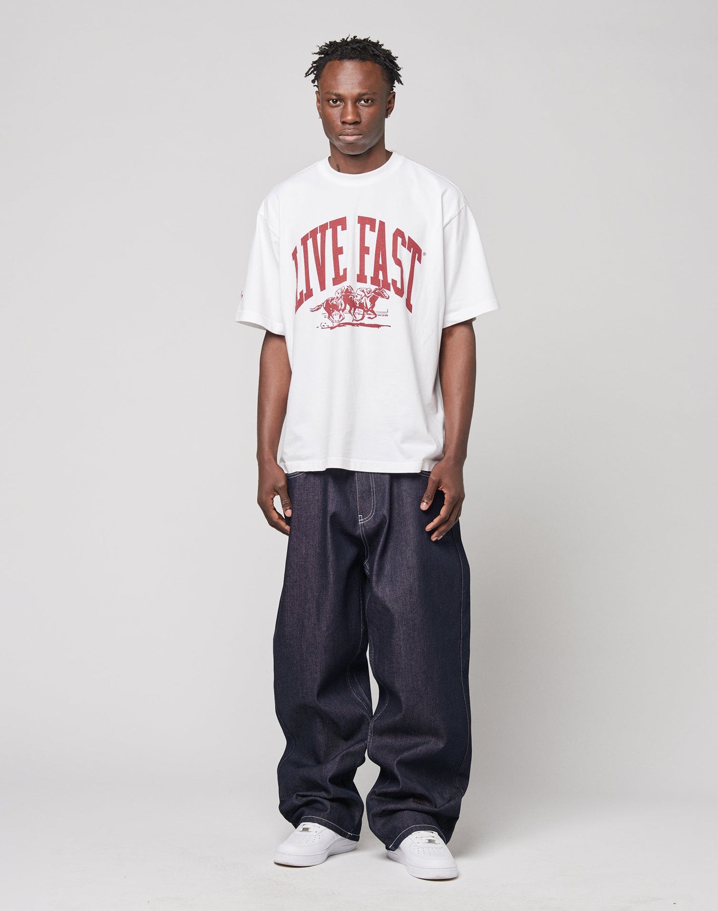 A man faces forward in a white Live Fast Stallion Tee by LIVE FAST DIE YOUNG, featuring an oversize fit and bold red LIVE FAST frontprint. He pairs it with dark baggy jeans and white sneakers. Made from soft Baumwolle.