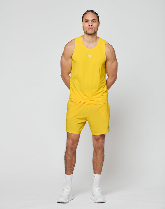 A man stands facing forward against a plain background, wearing LIVE FAST DIE YOUNGs bright yellow Live Fast Performance Shorts with Elastikbund and Reflektierender Print, paired with a matching tank top, white socks, and white sneakers. Hands behind his back.