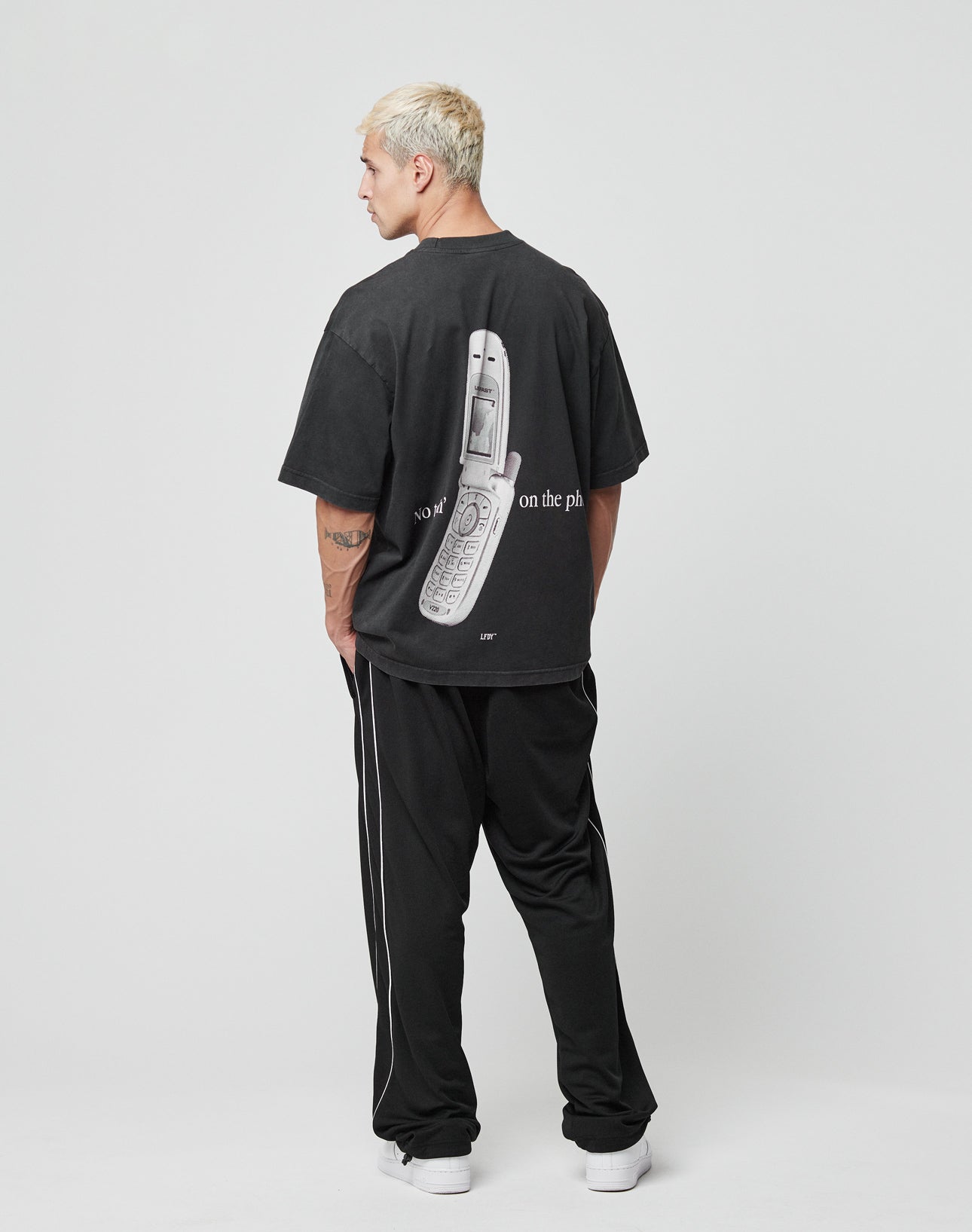 A man with short blond hair faces away, wearing the LIVE FAST DIE YOUNG LFDY Flip Phone Tee with a large flip phone graphic on the back, paired with black track pants with white stripes and white sneakers against a plain light background.
