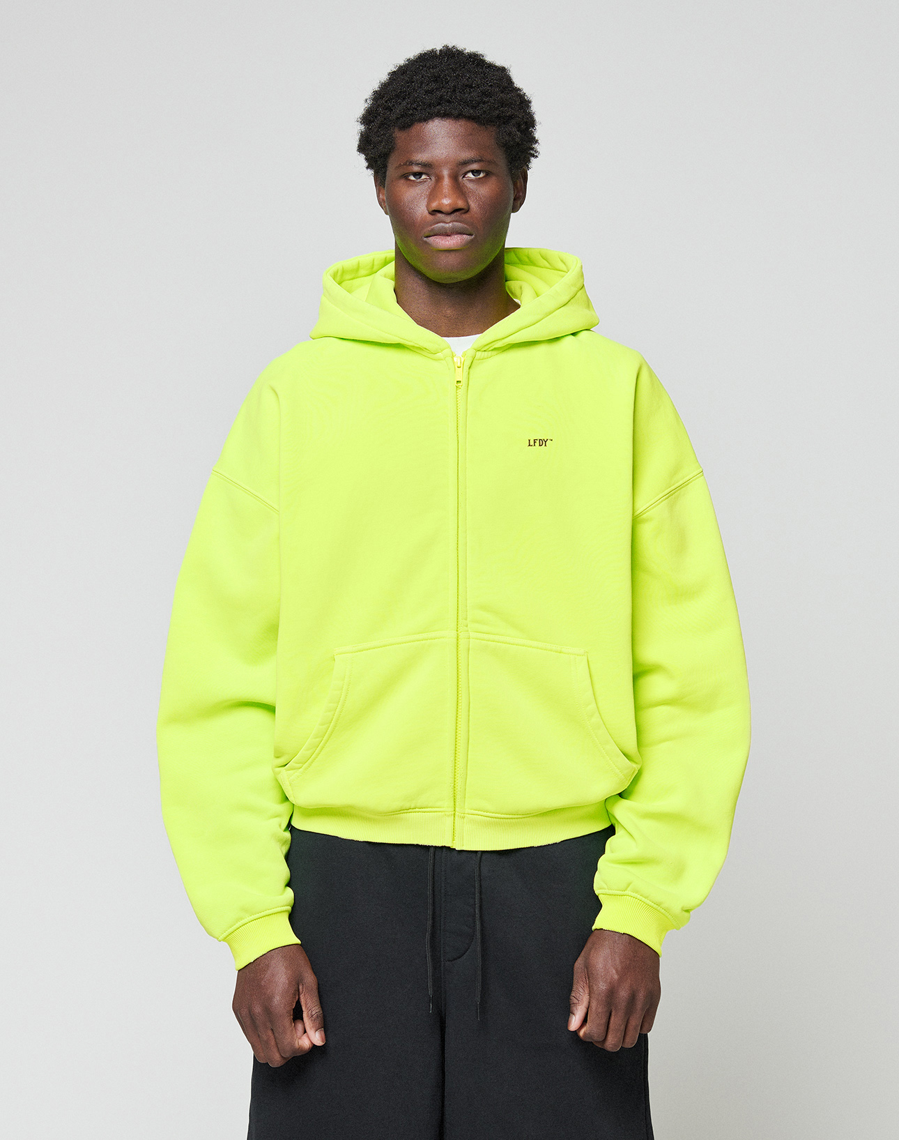 A person stands against a plain light gray background wearing the LIVE FAST DIE YOUNG Basic Hooded Zip in bright neon yellow with black pants. The zip-up hoodie features an oversized fit, and they look straight at the camera with a neutral expression. | model