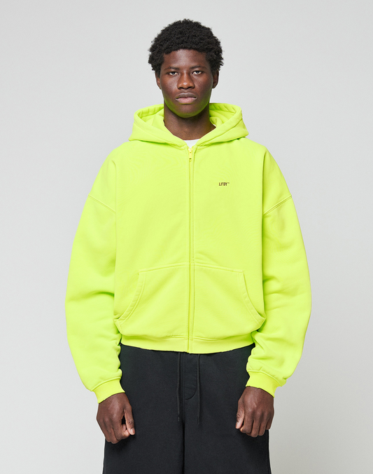 A person stands against a plain light gray background wearing the LIVE FAST DIE YOUNG Basic Hooded Zip in bright neon yellow with black pants. The zip-up hoodie features an oversized fit, and they look straight at the camera with a neutral expression. | model
