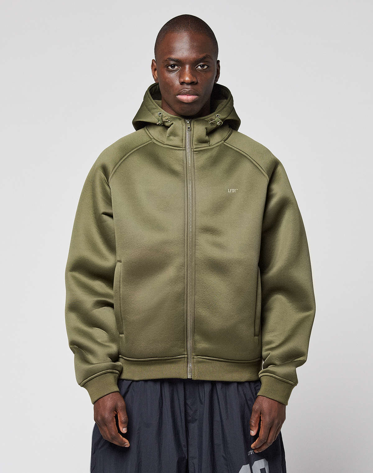 A person stands facing forward, wearing the LFDY Scuba Hooded Zip by LIVE FAST DIE YOUNG in olive green with a hood and zip pockets, regular fit, paired with dark gray pants against a plain light gray background. | model