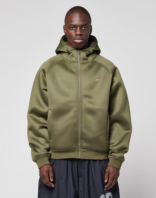 A person stands facing forward, wearing the LFDY Scuba Hooded Zip by LIVE FAST DIE YOUNG in olive green with a hood and zip pockets, regular fit, paired with dark gray pants against a plain light gray background. | model