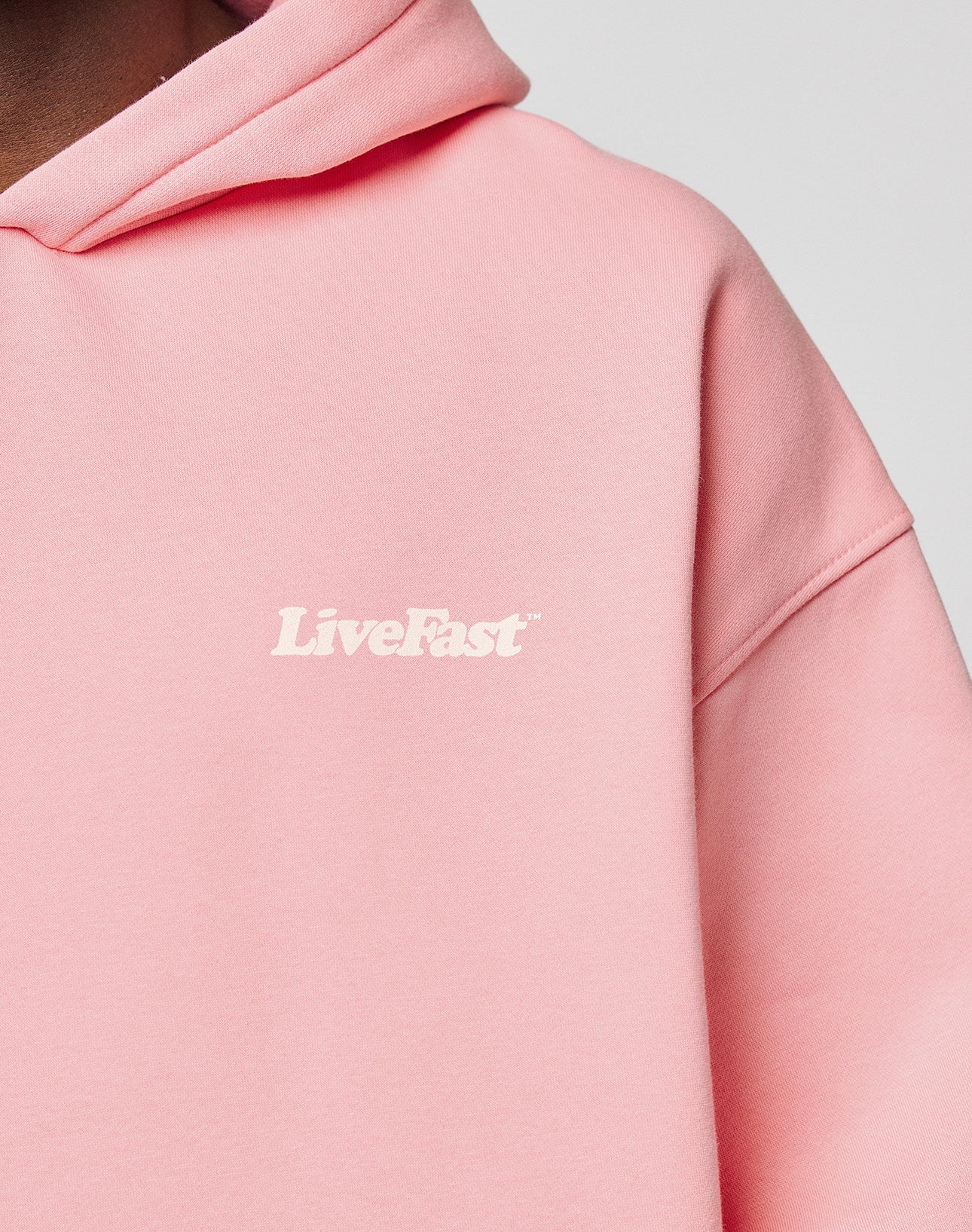 Close-up of a person wearing the LIVE FAST DIE YOUNG Basic 360 Hooded in light pink, featuring white LiveFast text on the left chest, topstitching details, and an oversize fit. Set against a plain, neutral background.