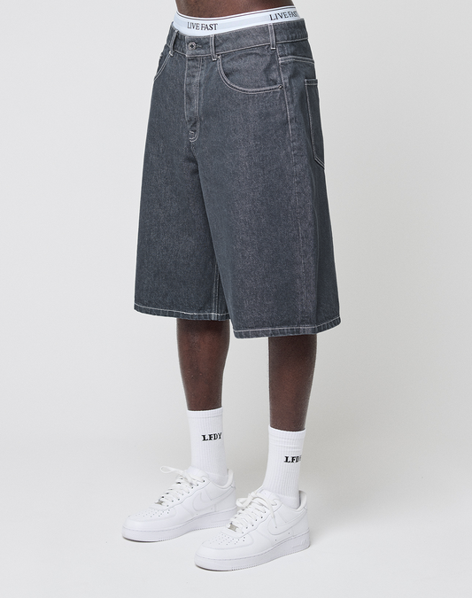 A person in baggy fit LFDY Jorts by LIVE FAST DIE YOUNG with a signature Lederpatch, white crew socks with black text, white sneakers, and white underwear featuring "LIVE FAST" on the waistband stands against a plain light background. | model