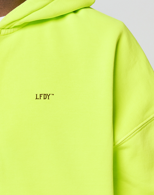 Close-up of the neon yellow LIVE FAST DIE YOUNG Basic Hooded Zip, featuring a small black LFDY™ logo on the chest. The image highlights the hood, partial sleeve, and signature branding on this oversized fit.