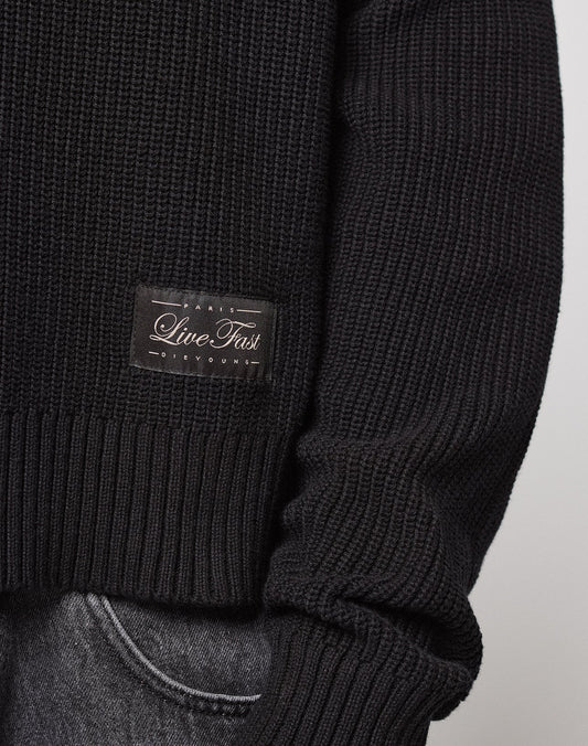 A close-up of a person wearing the LFDY Knit Sweater by LIVE FAST DIE YOUNG, featuring a dropped shoulder and a "Paris Live Fast" patch, styled with gray pants. The oversize fit is shown by the slightly rolled-up sleeve.