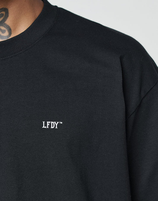 Close-up of a person wearing the LIVE FAST DIE YOUNG Basic Chest Tee, a Regular Fit black shirt with a white LFDY™ logo on the chest. Made from 100% Baumwolle for comfort. Their face is out of frame; part of a neck tattoo is visible.