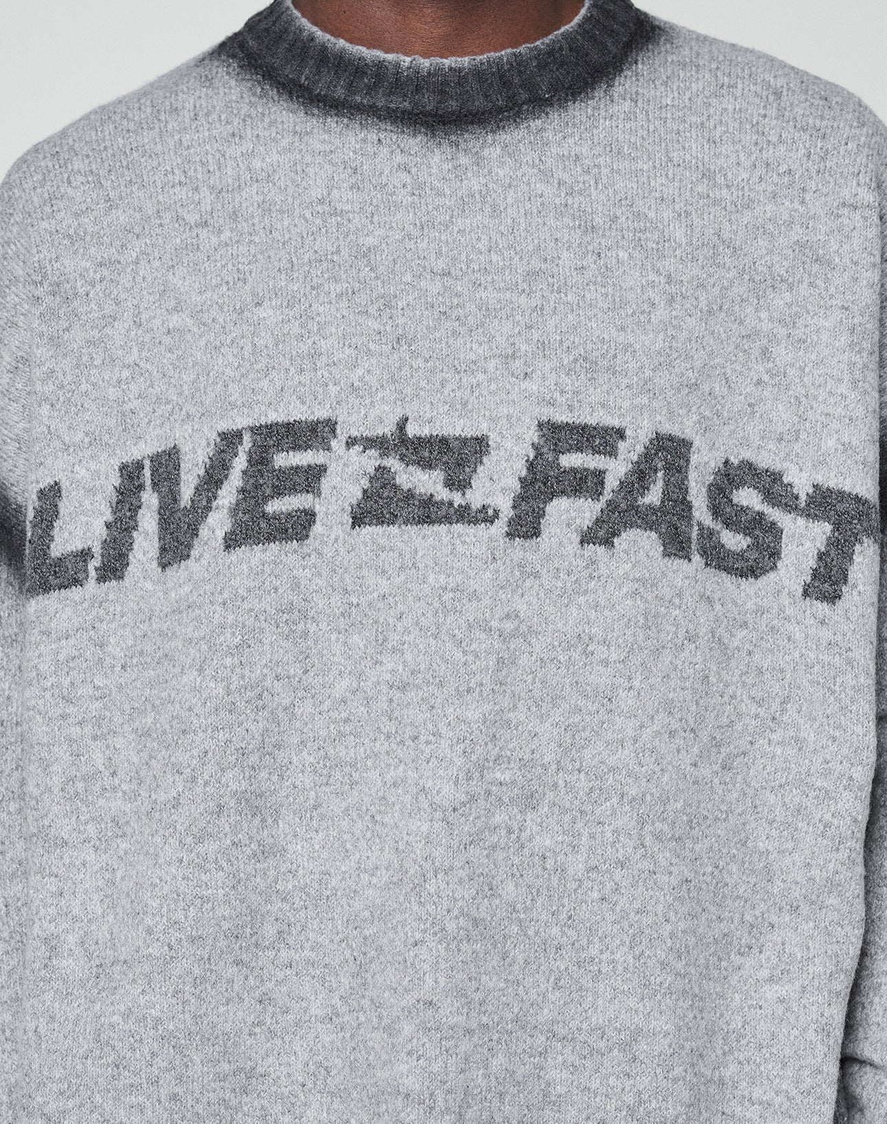 A close-up of the Live Fast Boucle Sweater by LIVE FAST DIE YOUNG, light gray with dark trim, features an oversized fit and bold, distressed LIVE FAST lettering on the chest, finished with a subtle faded spray effect.