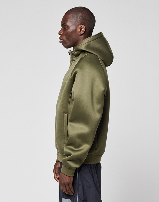 A person stands in profile facing left, wearing an olive green LFDY Scuba Hooded Zip by LIVE FAST DIE YOUNG with dark pants, set against a plain light gray background.
