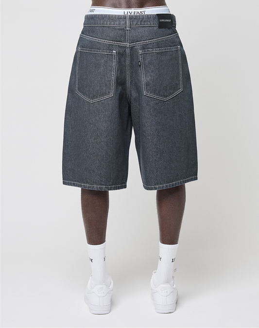 A person, back to the camera against a plain white background, wears baggy fit LIVE FAST DIE YOUNG LFDY Jorts with a leather patch, white LIVFAST socks, and white sneakers.