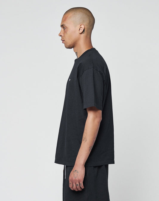 A person with a shaved head and tattoos stands in profile, wearing the LIVE FAST DIE YOUNG Basic Chest Tee— a loose black 100% Baumwolle T-shirt with subtle logo print— and black pants, set against a plain white background.