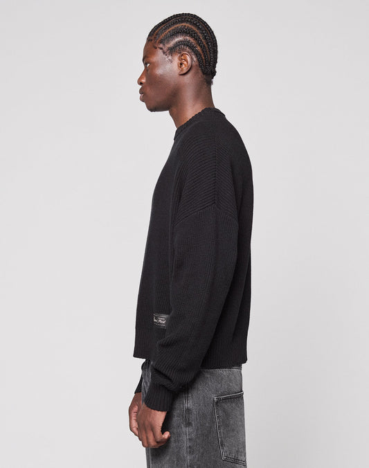 A person with braided hair wears the LIVE FAST DIE YOUNG LFDY Knit Sweater in black and washed black jeans, standing in profile against a plain light gray background.