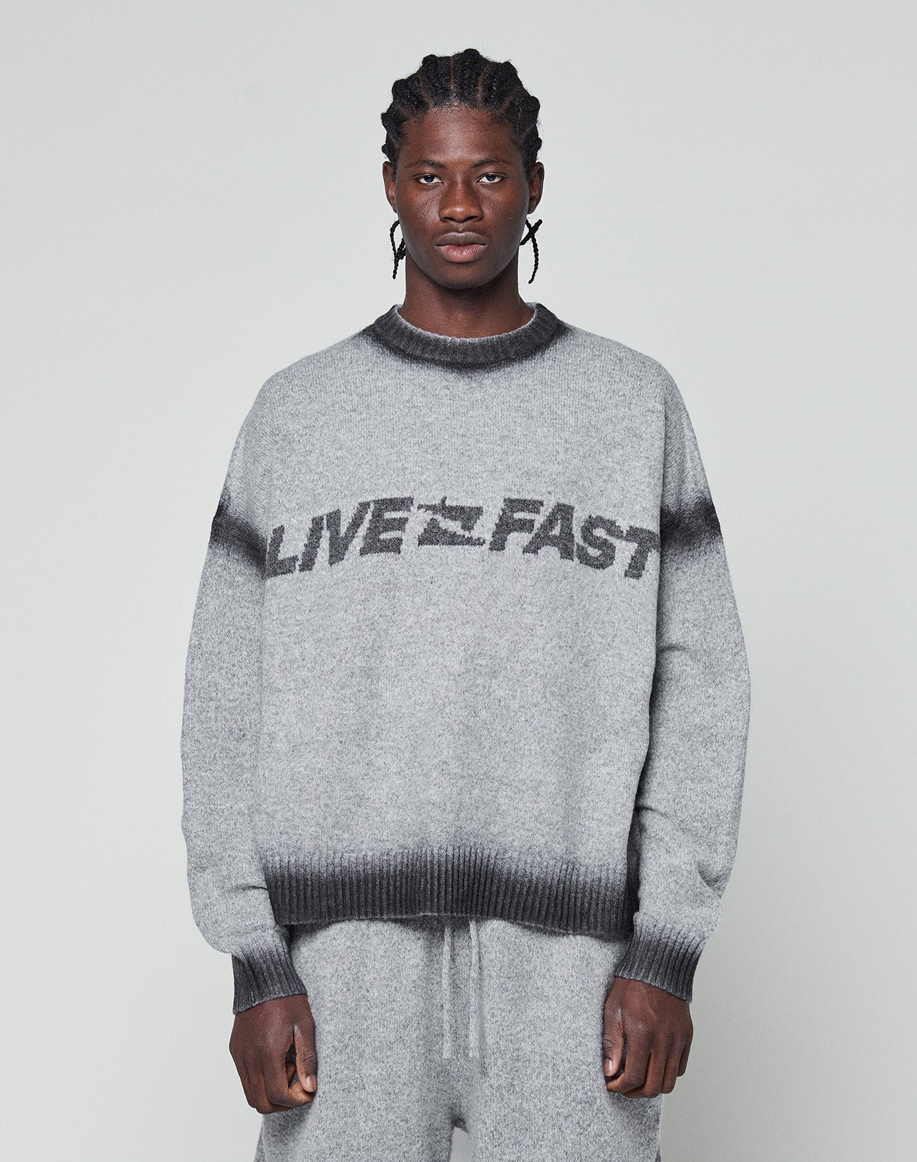 A man with braided hair wears an oversize fit grey Live Fast Boucle Sweater by LIVE FAST DIE YOUNG, featuring a dark faded spray effect stripe and bold LIVE FAST lettering, paired with matching pants against a plain light background.