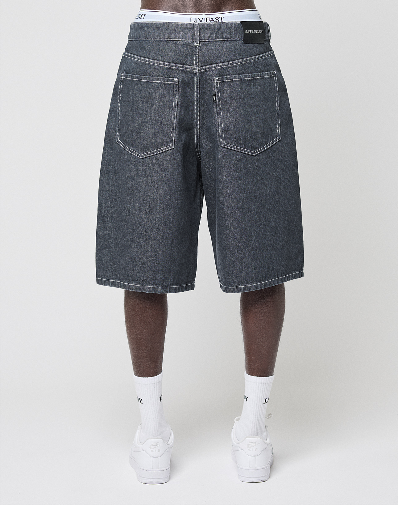 A person, back to the camera against a plain white background, wears baggy fit LIVE FAST DIE YOUNG LFDY Jorts with a leather patch, white LIVFAST socks, and white sneakers.