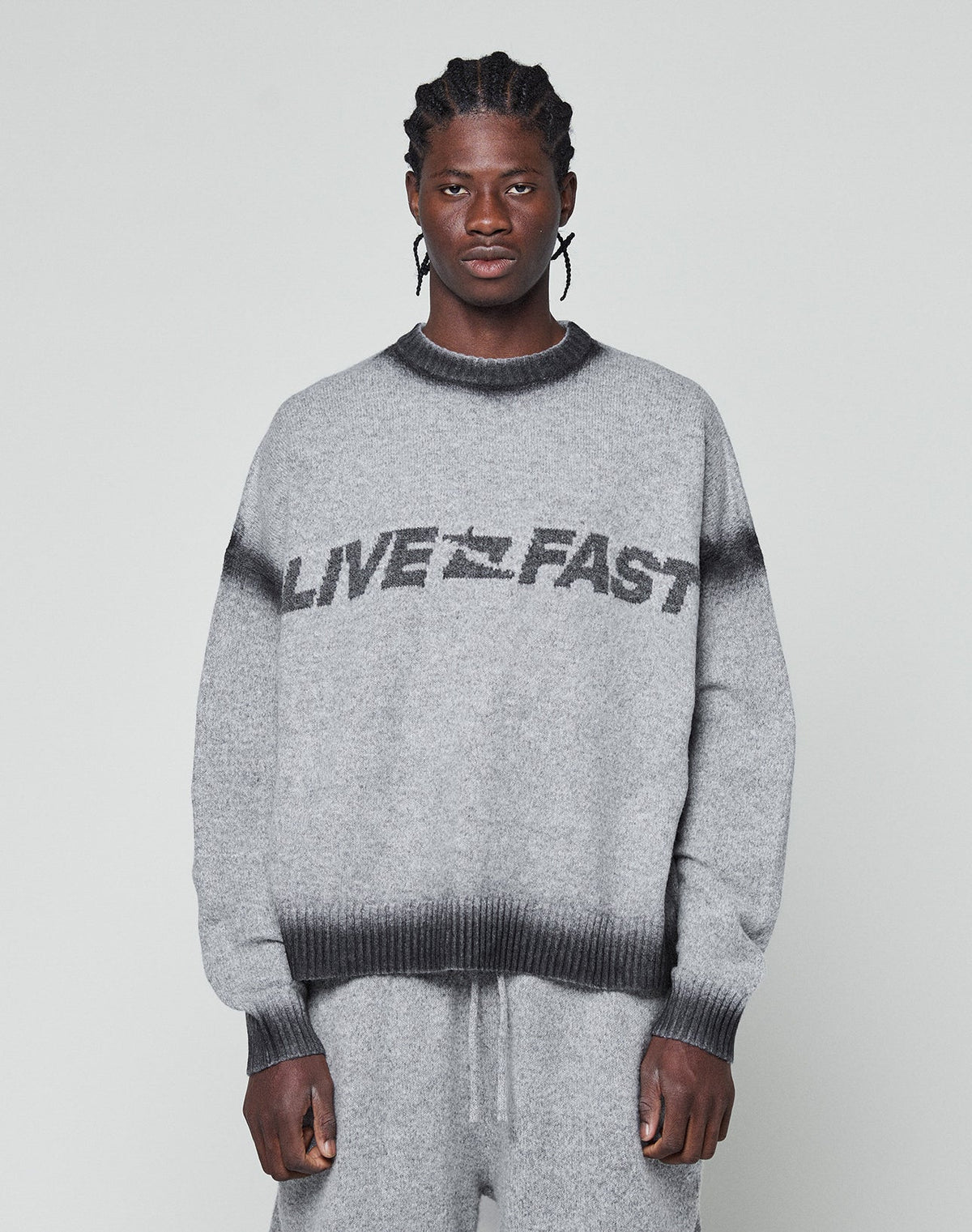 A man with braided hair wears an oversize fit grey Live Fast Boucle Sweater by LIVE FAST DIE YOUNG, featuring a dark faded spray effect stripe and bold LIVE FAST lettering, paired with matching pants against a plain light background. | model