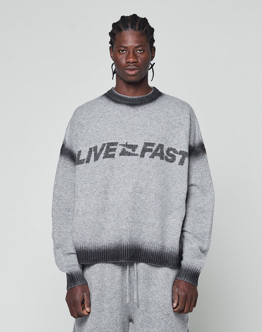 A man with braided hair wears an oversize fit grey Live Fast Boucle Sweater by LIVE FAST DIE YOUNG, featuring a dark faded spray effect stripe and bold LIVE FAST lettering, paired with matching pants against a plain light background. | model