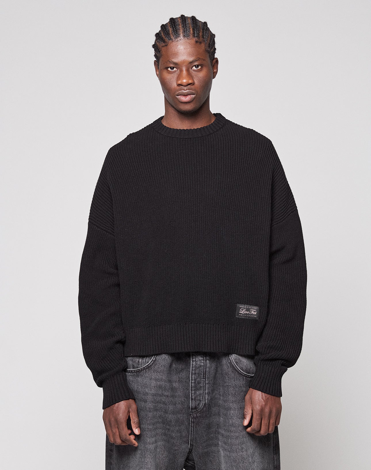 Wearing the LFDY Knit Sweater from LIVE FAST DIE YOUNG and baggy dark jeans, a person with braided hair stands against a plain light gray background, looking directly at the camera with a neutral expression.