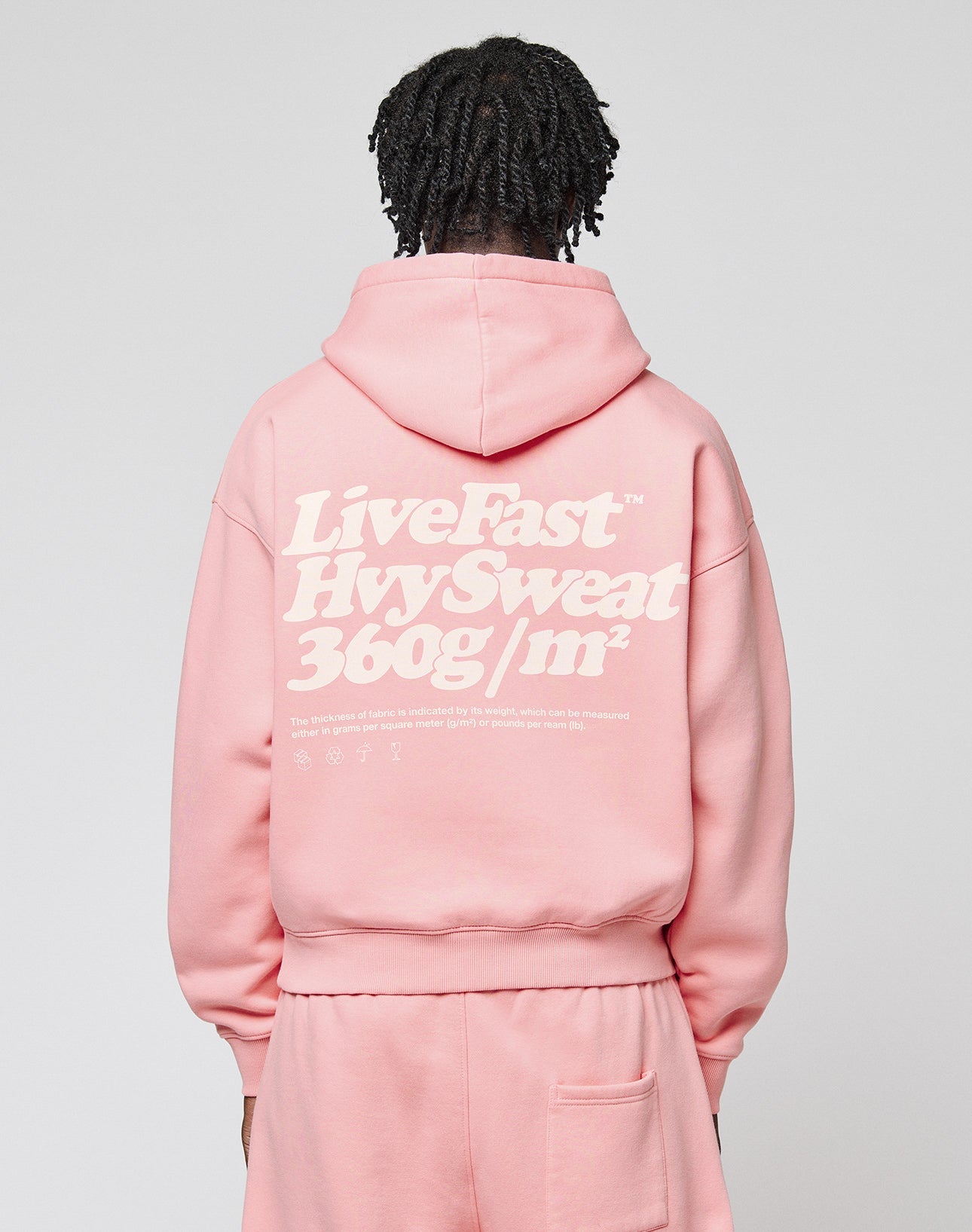 A person with short, twisted hair wears a pink LIVE FAST DIE YOUNG Basic 360 Hooded and matching pants. The hoodie has bold front and back print, sturdy topstitching, and white text. The background is plain.