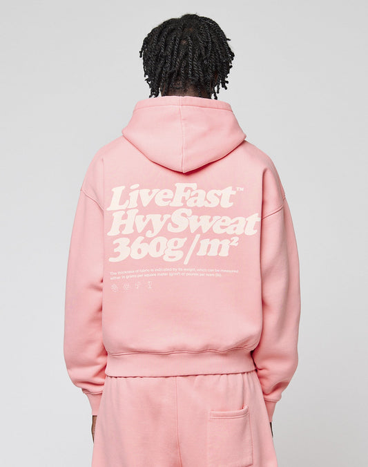 A person with short, twisted hair wears a pink LIVE FAST DIE YOUNG Basic 360 Hooded and matching pants. The hoodie has bold front and back print, sturdy topstitching, and white text. The background is plain.