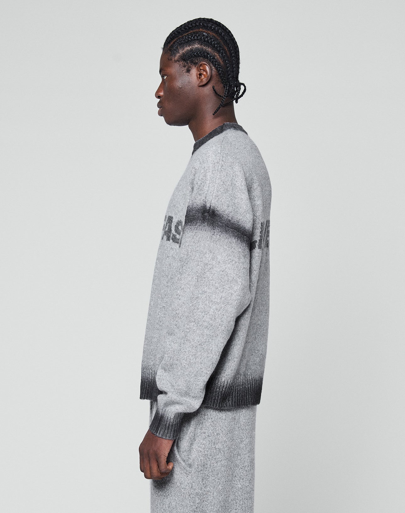 A person with braided hair stands in profile wearing the LIVE FAST DIE YOUNG Live Fast Boucle Sweater in gray, featuring an oversize fit. The matching pants have a dark horizontal stripe and subtle pattern. Background is plain light gray.