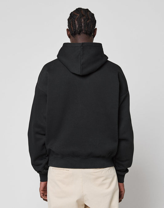 A person with braided hair is seen from behind in a LIVE FAST DIE YOUNG Monaco Boys Hooded oversize black sweatshirt (Baumwolle-Polyester mix) and light beige pants, standing against a plain light gray background.