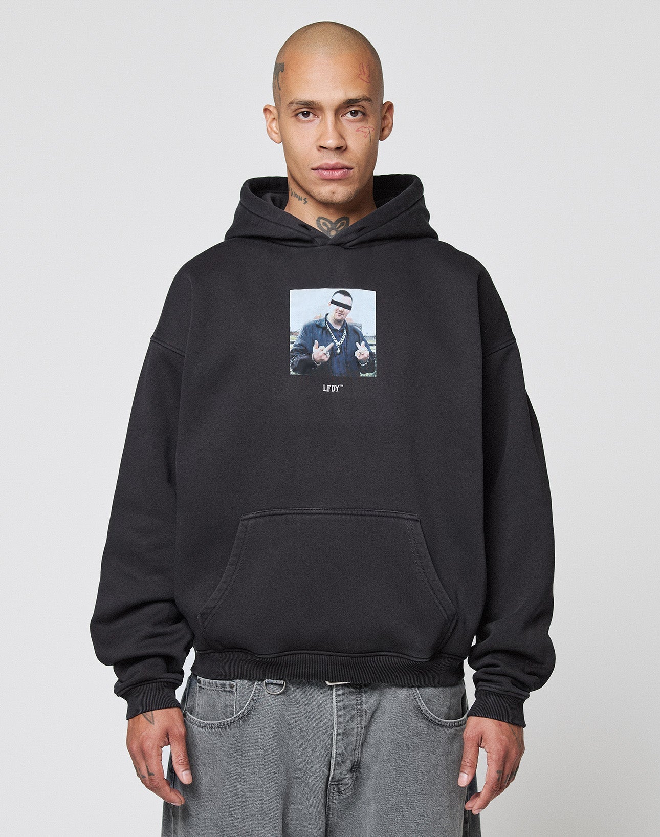 A person with a shaved head and neck tattoo stands facing forward, wearing the LIVE FAST DIE YOUNG LFDY Gambling Hooded black hoodie with chest frontprint and gray jeans against a plain light background.