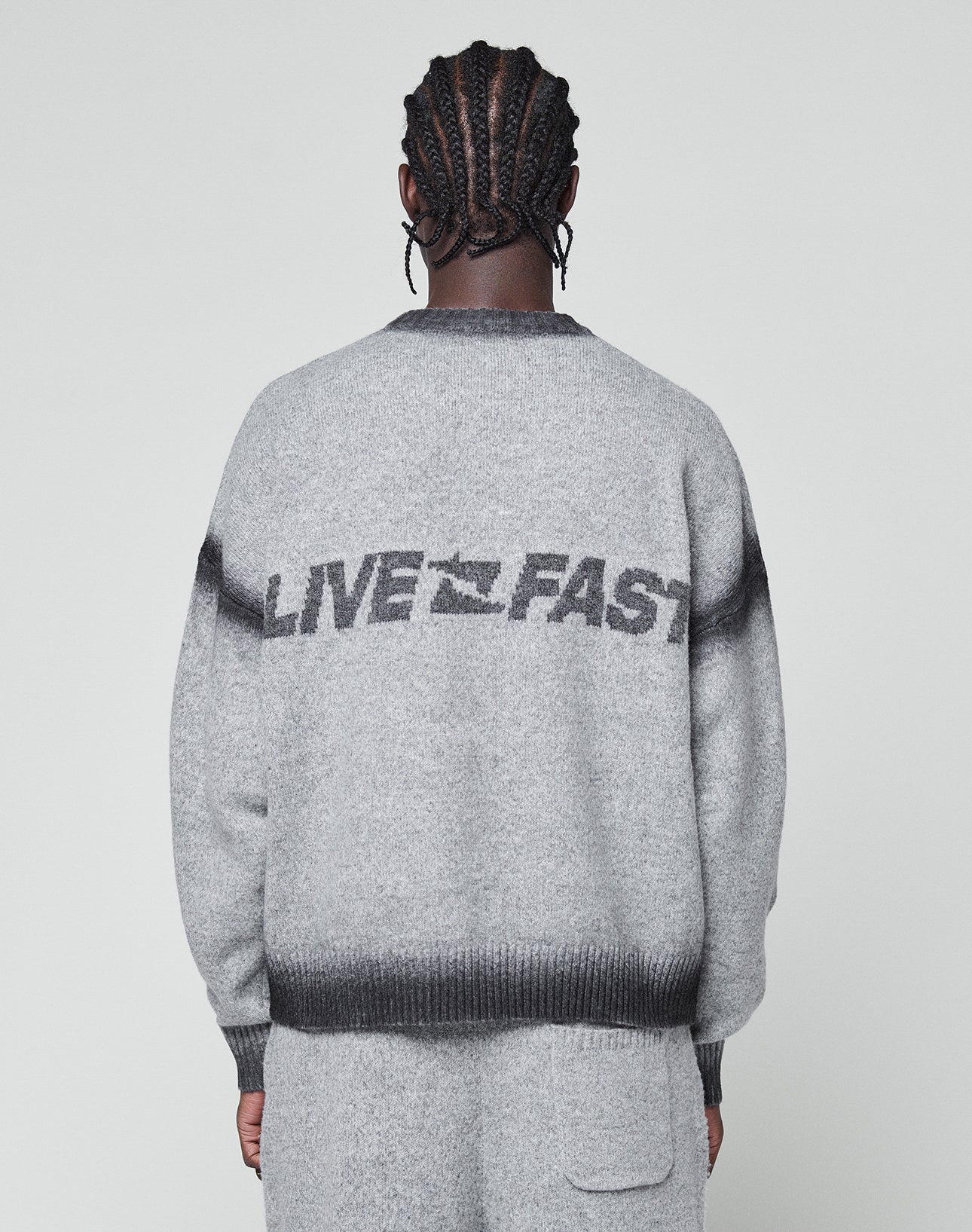 A person with braided hair, seen from behind, wears the LIVE FAST DIE YOUNG Live Fast Boucle Sweater in light gray. "LIVE FAST" and a bird graphic are on the back with a faded spray effect and dark gray stripes at shoulders and hem.