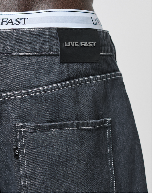 Close-up of someone wearing dark denim LFDY Jorts by LIVE FAST DIE YOUNG, featuring white stitching, a black LIVE FAST waistband label, and a visible white waistband above the classic Baggy Fit jeans.