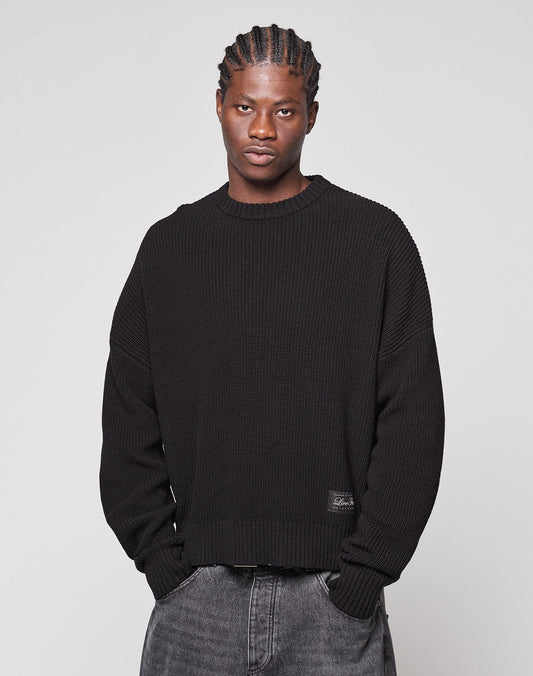A person with braided hair wears the LFDY Knit Sweater by LIVE FAST DIE YOUNG—an oversized, loose black 100% Baumwolle (cotton) knit with dropped shoulders—paired with faded gray jeans, standing against a plain light gray background.