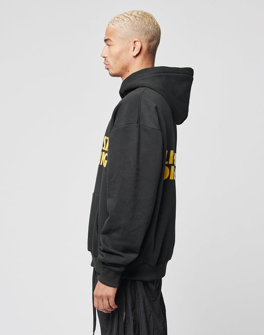 A man with short, bleached blond hair stands in profile wearing the LIVE FAST DIE YOUNG LFDY Drift Hooded, featuring a doppellagige Kapuze, yellow text, and an oversize fit with black pants against a plain light gray background.