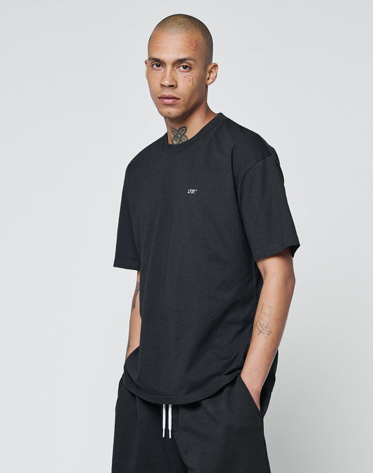 A person with a shaved head, facial and neck tattoos wears the LIVE FAST DIE YOUNG Basic Chest Tee in black, made from 100% Baumwolle, and black pants, standing hands-in-pockets against a plain light gray background.