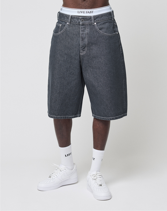 A person wearing baggy-fit dark denim LFDY Jorts by LIVE FAST DIE YOUNG, featuring a white LIVE FAST waistband, with white crew socks and sneakers, stands against a plain light background.