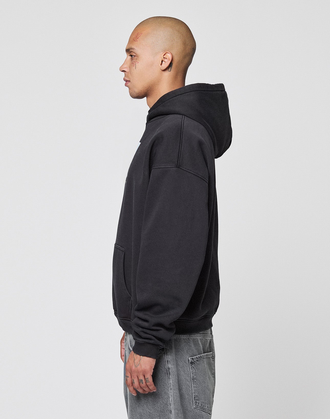 A tattooed person with a shaved head stands in profile facing left, wearing an oversize fit LIVE FAST DIE YOUNG LFDY Gambling Hooded in black and faded gray jeans against a plain light gray background.