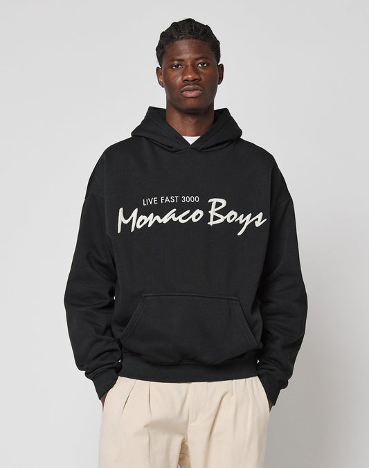 A man in an oversized black Monaco Boys Hooded by LIVE FAST DIE YOUNG stands against a light gray background. He has short hair and wears light-colored pants made of a soft cotton-polyester blend.