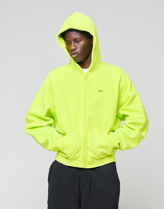 A person in a neon yellow LIVE FAST DIE YOUNG Basic Hooded Zip with hood up and black pants stands against a plain light gray background, looking down with hands in pockets.