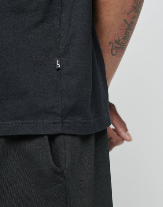 A close-up shows a tattooed person in a black LIVE FAST DIE YOUNG Basic Chest Tee and black pants, one hand in the pocket. A small black tag with white text on the side seam highlights the subtle Logo Print detail.