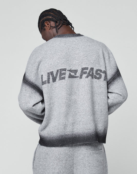 A person with braided hair faces away, wearing the LIVE FAST DIE YOUNG Live Fast Boucle Sweater—an oversize gray piece featuring bold “LIVE FAST” lettering on the back. The background is plain and light-colored.