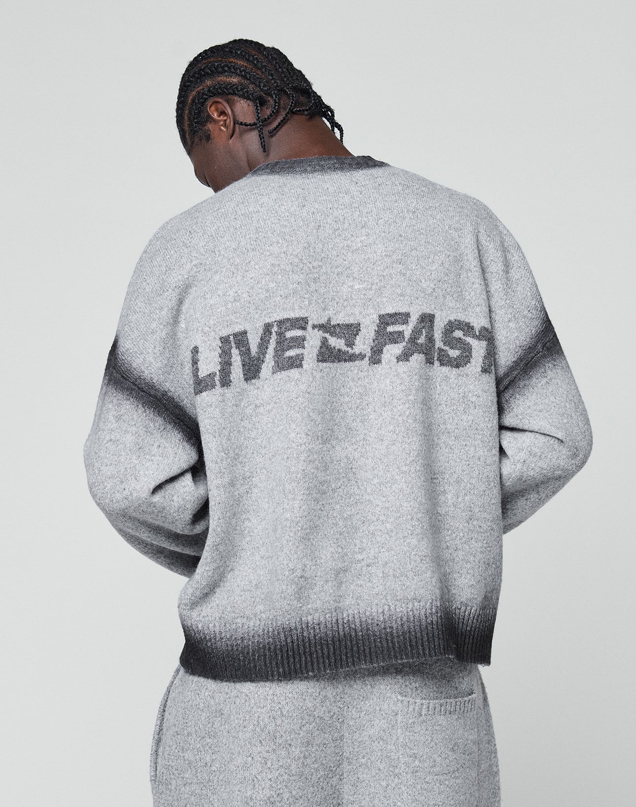 A person with braided hair faces away, wearing the LIVE FAST DIE YOUNG Live Fast Boucle Sweater—an oversize gray piece featuring bold “LIVE FAST” lettering on the back. The background is plain and light-colored.