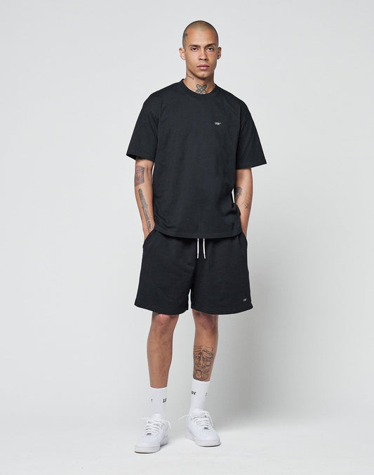 A person stands against a white background wearing the LIVE FAST DIE YOUNG Basic Chest Tee in black, paired with black shorts, white crew socks, and white sneakers. Their hands are in their pockets, showing tattoos on their arms and legs.
