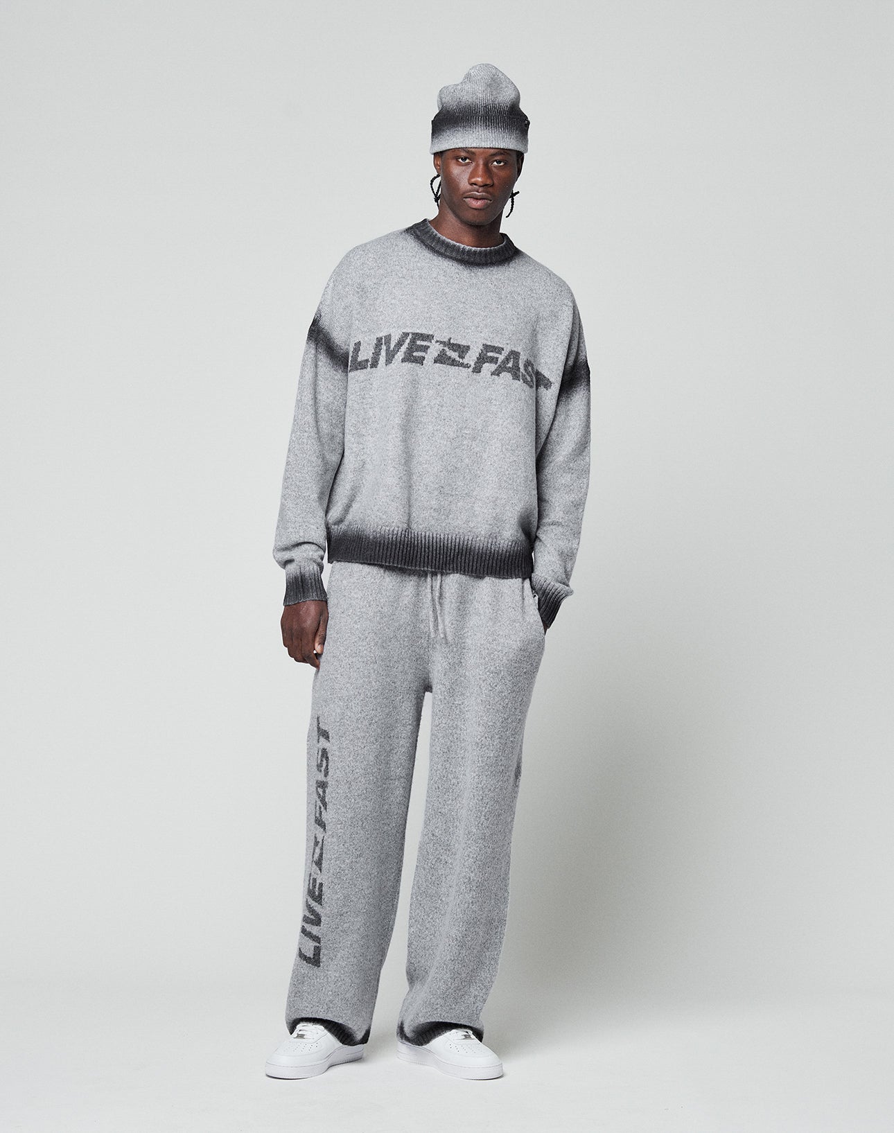 A person wearing the LIVE FAST DIE YOUNG Live Fast Boucle Sweater and matching gray pants with "LIVE FAST" text stands against a light background, hands in pockets, finishing the look with a gray knit beanie and white sneakers.