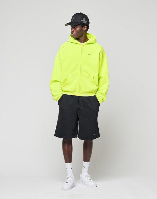 A person in an oversize fit neon yellow Basic Hooded Zip by LIVE FAST DIE YOUNG, black shorts, white socks, white sneakers, and a black baseball cap stands against a plain background, looking to the side with hands in pockets.