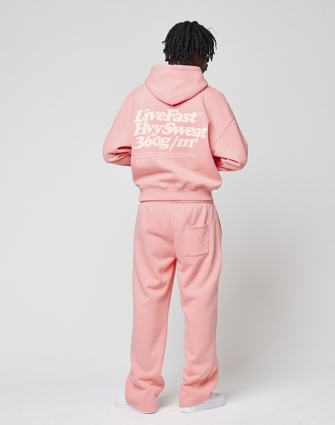 Someone stands facing away, wearing the LIVE FAST DIE YOUNG Basic 360 Hooded set in light pink with bold white Live Fast—Hvy Sweat 360g/m² back print. The oversized hoodie and sweatpants are shown against a plain white background.