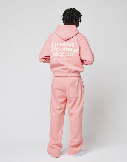 Someone stands facing away, wearing the LIVE FAST DIE YOUNG Basic 360 Hooded set in light pink with bold white Live Fast—Hvy Sweat 360g/m² back print. The oversized hoodie and sweatpants are shown against a plain white background.