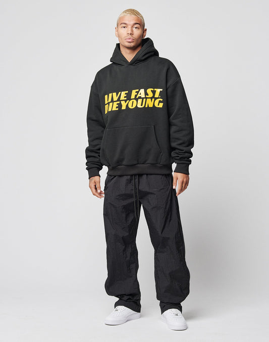 A person stands facing forward wearing the LFDY Drift Hooded by LIVE FAST DIE YOUNG, an oversize black hoodie with bold yellow LIVE FAST DIE YOUNG text, paired with black pants and white sneakers against a plain light gray background.