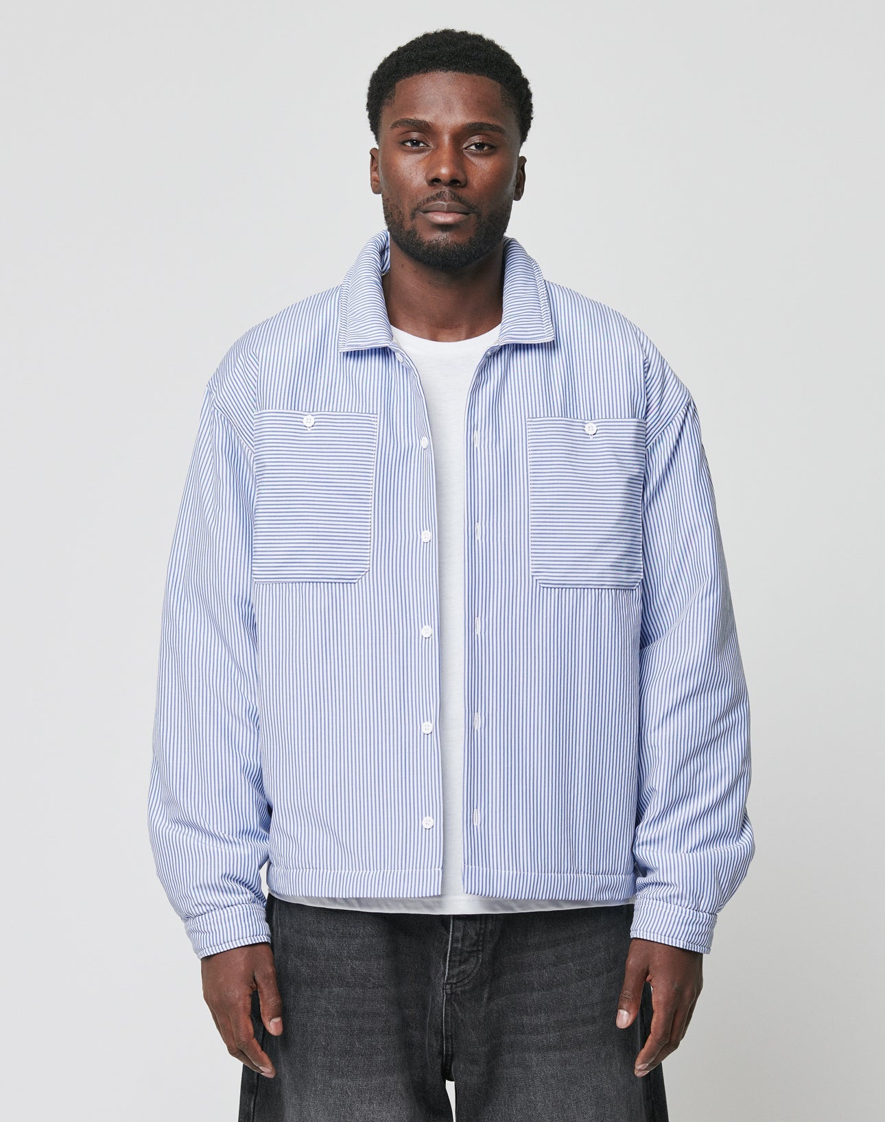 A man wearing the LIVE FAST DIE YOUNG Padded Protection Shirt—an oversized blue and white striped button-up—over a white tee and dark jeans stands facing the camera against a plain light gray background.