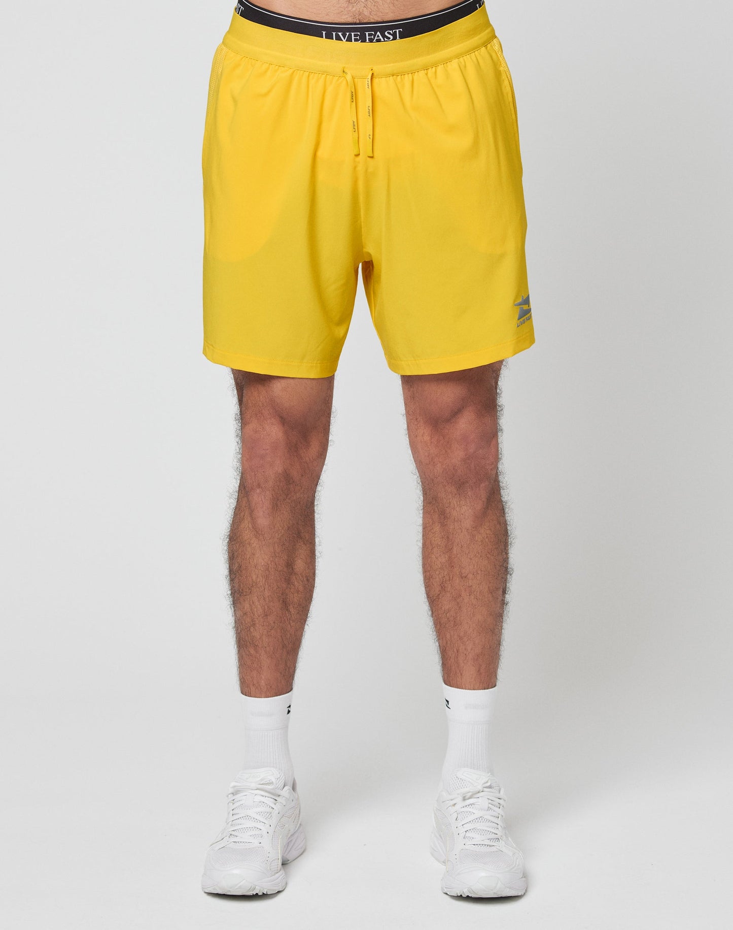 Someone stands against a white background wearing LIVE FAST DIE YOUNG’s bright yellow Live Fast Performance Shorts with a black waistband that reads LIVE FAST, paired with white socks and white sneakers.