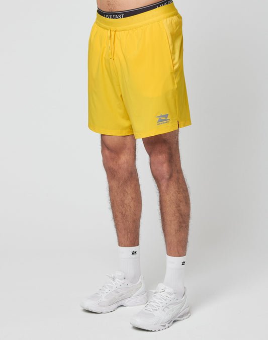 A person models the LIVE FAST DIE YOUNG Live Fast Performance Shorts in bright yellow with an elastic waistband and small reflective print near the hem, paired with white socks and athletic sneakers. The upper body is not shown.
