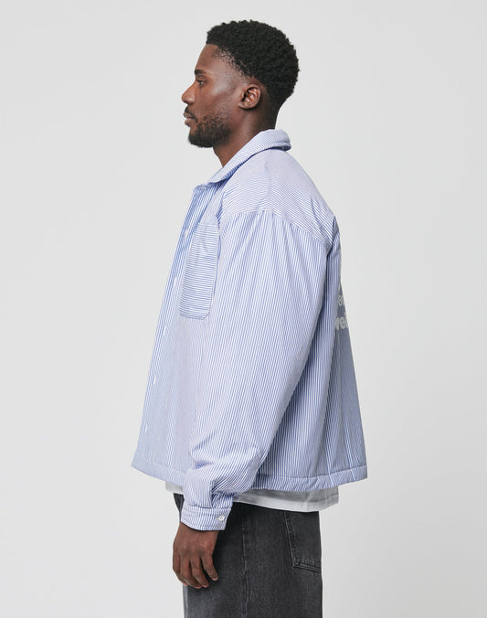 A man stands in profile against a plain background, wearing the LIVE FAST DIE YOUNG Padded Protection Shirt—an oversized blue and white vertically striped button-up with a chest pocket—paired with dark jeans.