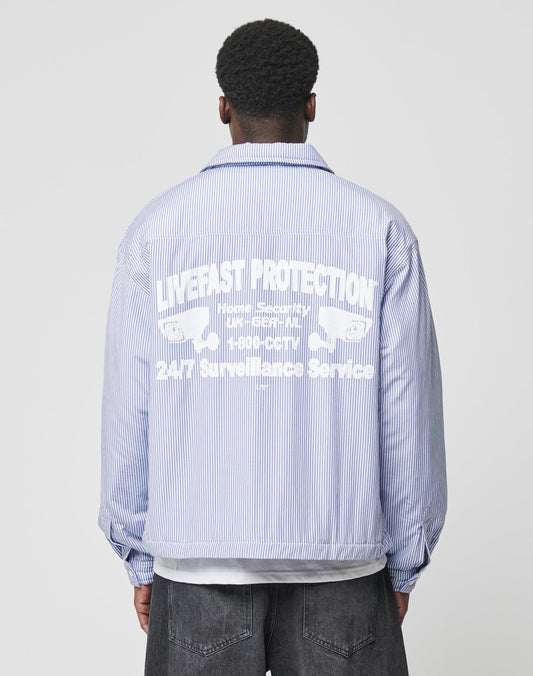 A person faces away, wearing the LIVE FAST DIE YOUNG Padded Protection Shirt—an oversized blue and white pinstriped jacket featuring a bold flag-label backprint with LIVEFAST PROTECTION, stylized text, and symmetrical graphics.